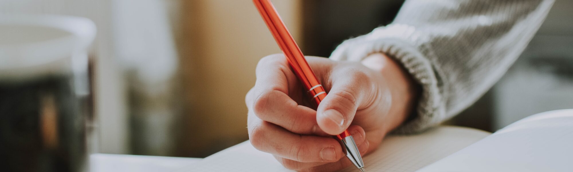 Eine Hand schreibt mit einem orangenen Stift in ein aufgeschlagenes Buch.