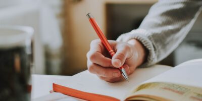 Eine Hand schreibt mit einem orangenen Stift in ein aufgeschlagenes Buch.
